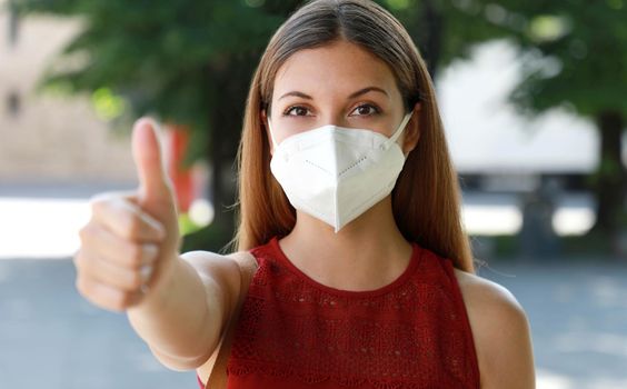 COVID-19 Optimistic girl wearing protective mask KN95 FFP2 avoiding Coronavirus disease 2019 showing thumbs up in city street