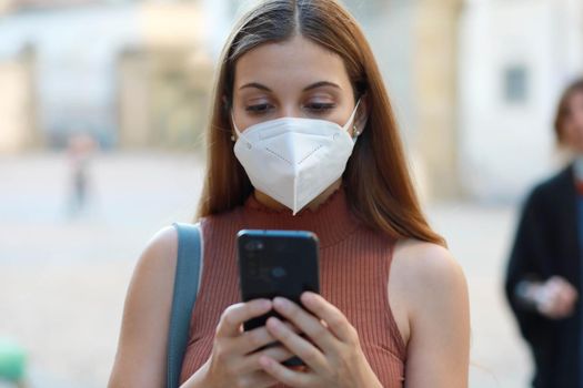 Portrait of young elegant woman wearing KN95 FFP2 mask messaging with mobile phone in city street