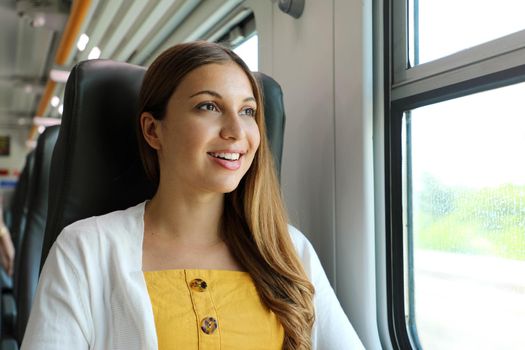 Portrait of young woman looking through the train window. Happy train passenger traveling sitting in a seat and looking through the window.