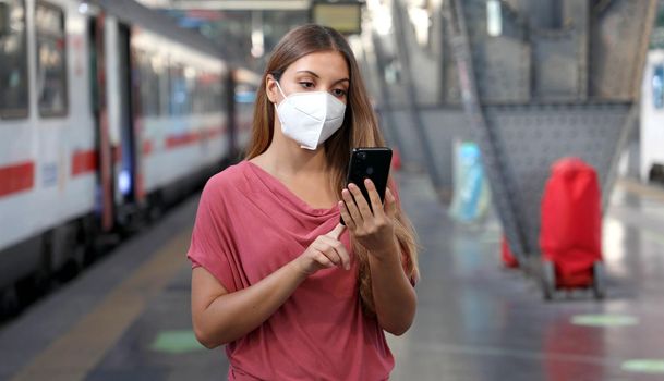 Young woman wearing KN95 FFP2 face mask waiting train at the station. Casual woman using smart phone in train station.