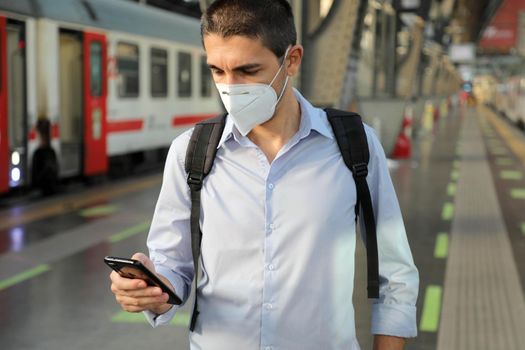 Business man wearing KN95 FFP2 face mask waiting train at the station. Young caucasian man using smart phone in train station.