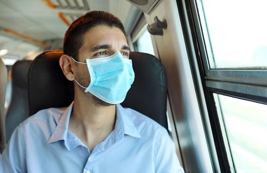 Travel safely on public transport. Young man with surgical mask looking through train window. Train passenger with protective mask looking through the window.
