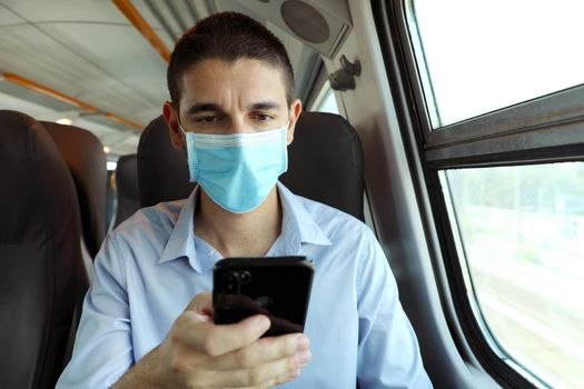 Man with surgical mask using smart phone app on public transport. Train passenger with protective mask traveling sitting in business class texting on mobile phone.