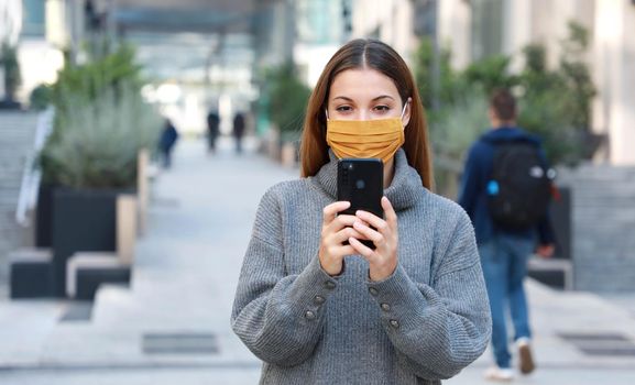 Young woman with fabric mask messaging with mobile phone while walking in pedestrian street. Copy space.
