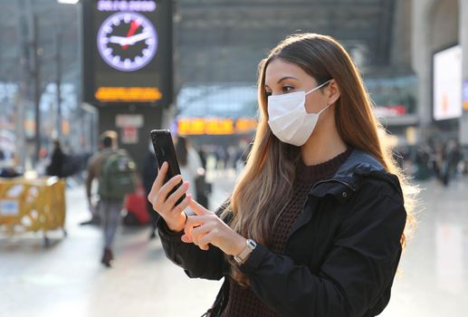 Business woman wearing protective mask buying ticket online with smartphone app at train station