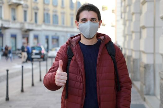 Portrait of optimistic young man wearing protective mask showing thumbs up in city street