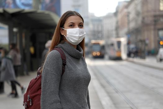 Student girl with protective mask waiting public transport in street. Young woman with surgical mask waiting tram at the stop in gray polluted city.