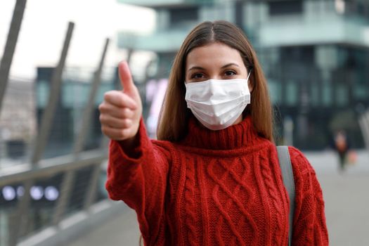 Optimistic young woman wearing protective mask showing thumbs up in city street ready for the vaccination campaign against Coronavirus disease 2019