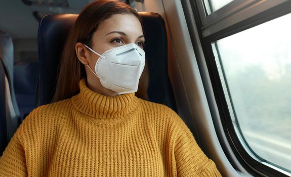 Travel safely on public transport. Young business woman with KN95 FFP2 face mask looking through train window. Train passenger with protective mask travels sitting in business class looking through the window.