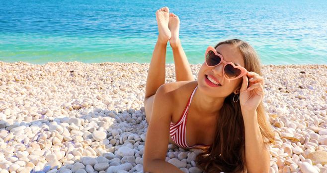 Sexy bikini girl lying on pebbles beach enjoying sun with heart sunglasses in her summer vacations. Copy space.