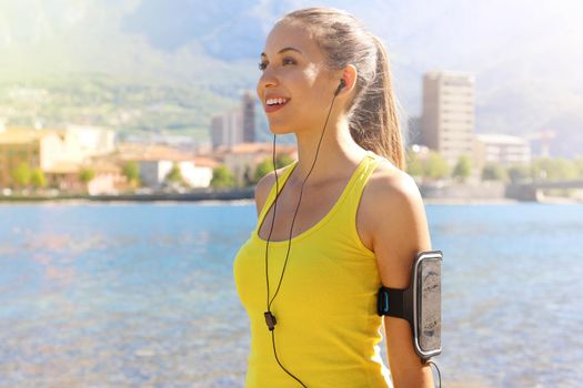Happy fitness woman living a fit healthy lifestyle. Young girl wearing activewear and sports armband for phone and earphones, tech gear for running or cardio workout city lake on background.