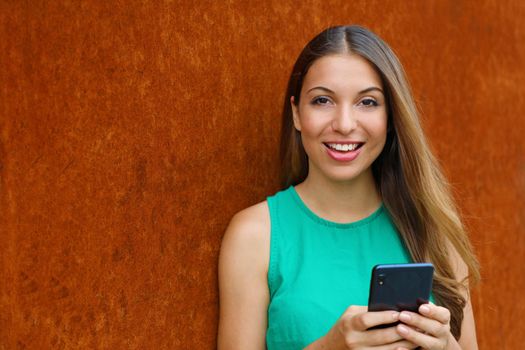 Young woman using smart phone looking at camera on rust background.