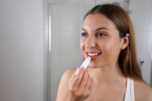 Smiling young woman applying lip balm in front of the mirror indoor