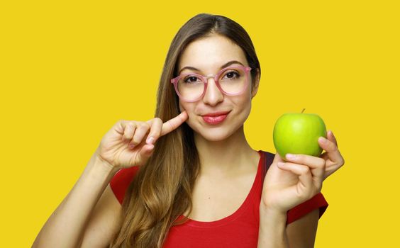 Portrait of a smiling young school nerd girl holding an apple gesturing like healthy good food isolated over yellow background