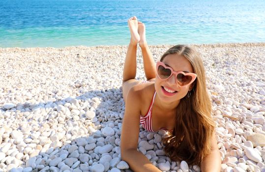 Portrait of young woman with heart sunglasses lying on pebbles beach. Summer holidays concept.
