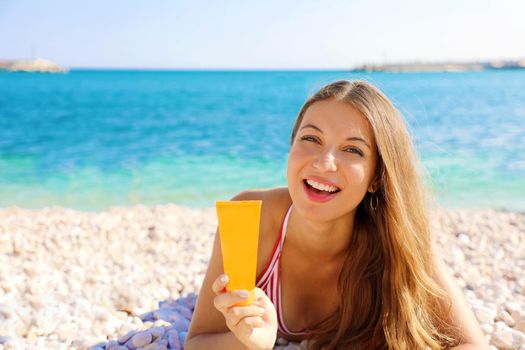Smiling woman holding sun cream tube protection lying on pebbles beach. Sunscreen girl showing suntan lotion in plastic container.