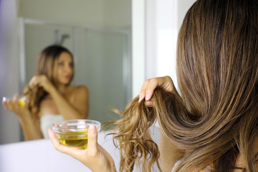 Young woman applying olive oil mask to hair tips in front of a mirror. Haircare concept. Focus on hair.