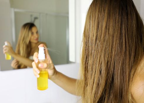 Young woman applying oil mask spray on hair tips in front of a mirror. Haircare concept. Focus on hair.
