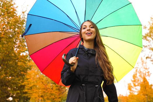 Pretty beautiful girl under rainbow rain umbrella in the park. Young woman looking at camera with colorful umbrella outdoor in Autumn season.