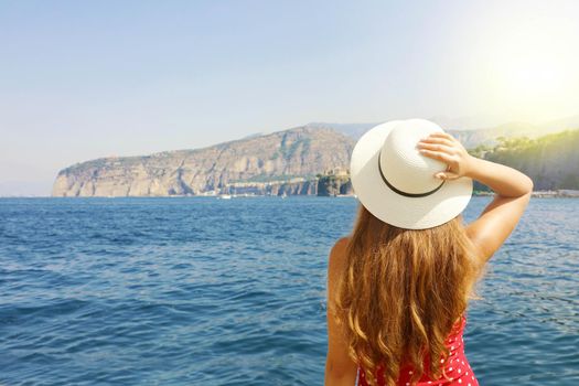 Summer holiday in Italy. Back view of young woman holding her hat with Sorrento Peninsula on the background, Sorrento Coast, Italy.