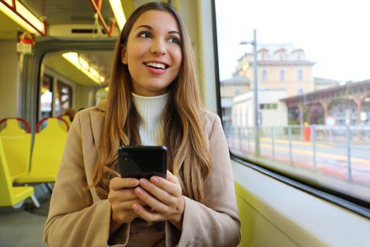 People on public transport. Smiling beautiful young woman holding mobile phone looking through the window on the tram.