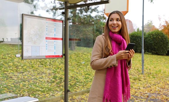 People and public transport. Smiling beautiful young woman holding mobile phone watch the bus arrive on bus stop.