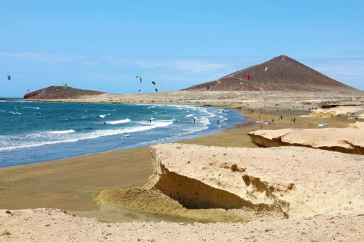 Many colorful kites on beach and kite surfers riding waves and flying during windy day in canarian El Medano in Tenerife with Montana Roja hills on horizon
