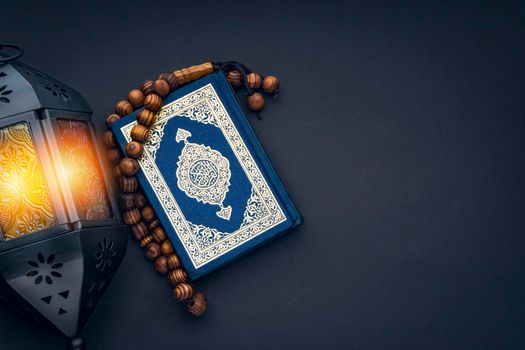 Holy Al Quran with written arabic calligraphy meaning of Al Quran, lantern lamp and rosary beads or tasbih on black background.