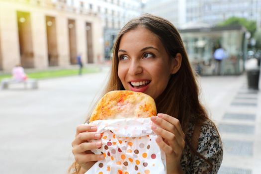 Happy young business woman eating a small pizza in her short lunch break. Urban girl eating traditional italian pizza in Milan main street, Italy. Pizza lovers concept.