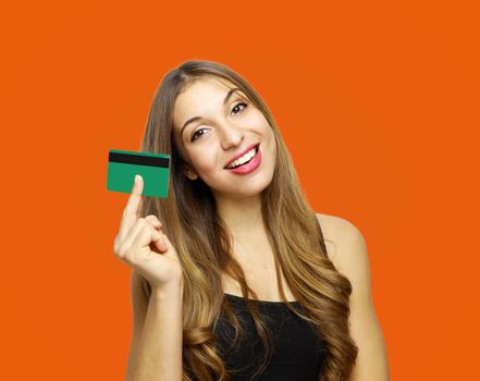 Image of smiling young lady standing over orange wall and holding debit card in hands. Looking at camera.