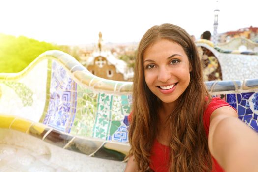 Smiling young woman tourist taking selfie self portrait sitting on the bench decorated with mosaic in famous Park Guell, Barcelona