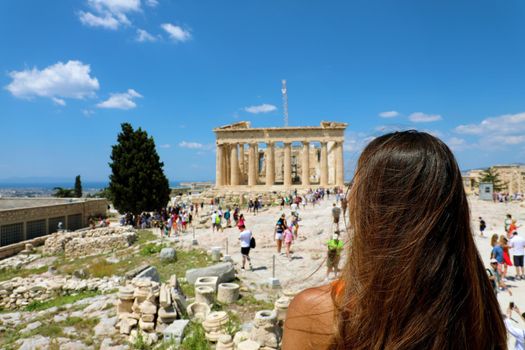 Young woman looks at Parthenon on the Acropolis of Athens, Greece. The famous ancient Greek Parthenon is the main tourist attraction of Athens. Girl traveler visits the Parthenon at the top of hill.