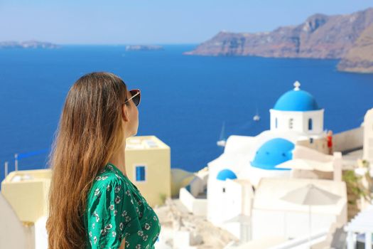 Santorini travel tourist woman visiting Oia, famous white village with blue domes in Greece. Girl in green dress and sunglasses looking to famous blue dome churches. European destination