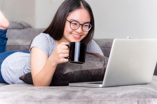 Portrait beautiful Asian young woman lying relax hold coffee cup work with a laptop, Cute girl teens looking at monitor smile happily on the sofa working remotely with an internet computer from home