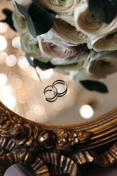 Wedding rings on a mirror surface with a bouquet of flowers.
