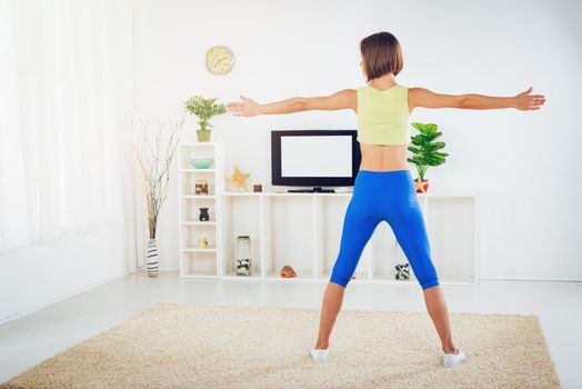 Rear view of an fit woman warming up while doing stretching exercises at home in front TV.