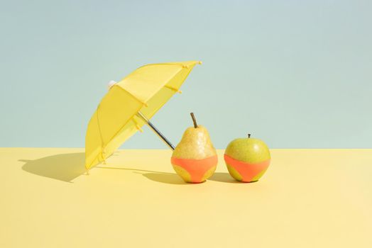 Summer fruit concept. Wet apple and a pear in monokini next to an umbrella isolated on a blue and yellow background.