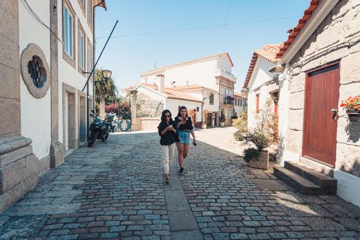 Two women walking through the streets of a ancient portuguese villa while looking their phones