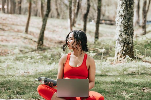 Young woman in fitness clothes relaxing on his laptop grabbing a bottle of water at the park over a yoga mat