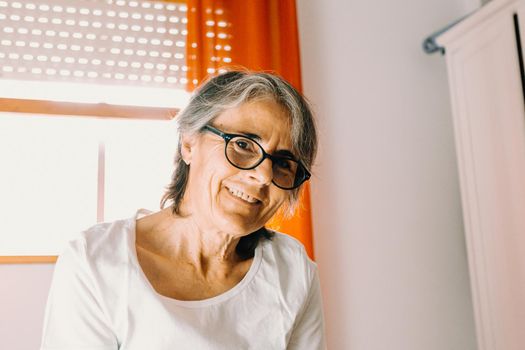 Close up of a old woman with glasses smiling to camera in a bright bedroom