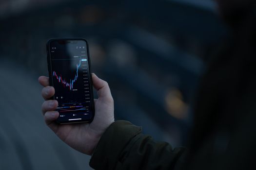 Male investor using stock trading app on smartphone, trader analyzing dynamic on forex chart and price flow on screen while standing outdoors. Selective focus on hand holding mobile phone