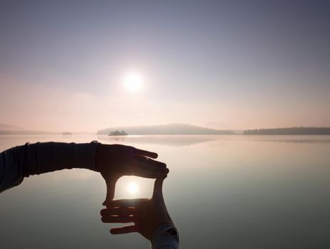 Man hand makes shadow symbols  to Sun. Misty daybreak at lake.