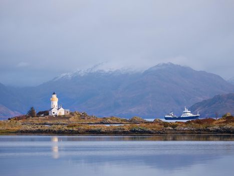 Lighthouse on Isle of Ornsay.Trade ship at rocky island, mountains in background. 