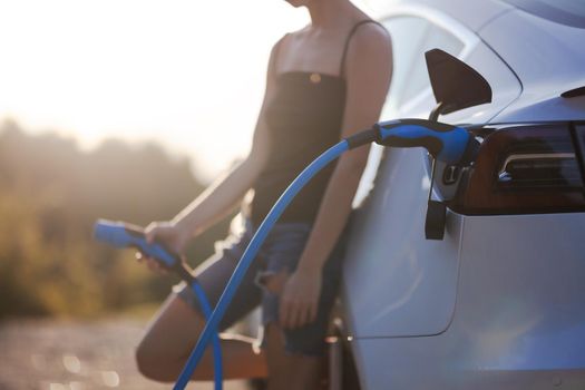 Beautiful young girl next to an electric car. Holding a charging cable. Sunset backlight