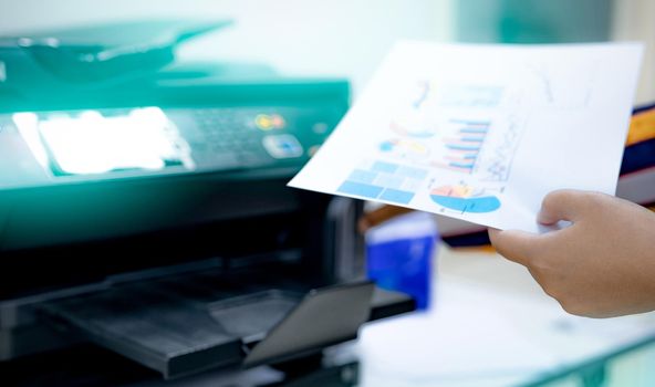 Selective focus on office worker hand holding paper. Multifunction laser printer. Copy, print, scan, and fax machine in office. Modern print technology. Photocopy machine. Document and paper work. 