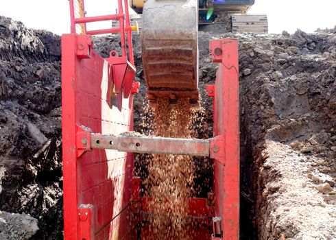 excavator filling dip excavation supported by trench box with pipe bedding pea gravel during installation of drainage pipe