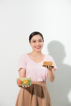 Half-length portrait of very beautiful woman holding small cake, fresh vegetables. Young housewife choosing sweets or healthy eating - cake and salad. Isolated on white background.