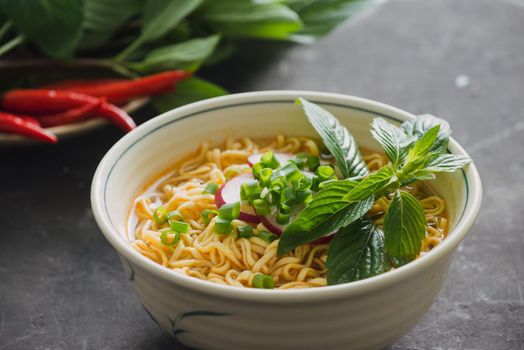 Instant noodles in bowl with fresh herbs, garnish of cilantro and Asian basil, lemon, lime on dark stone background