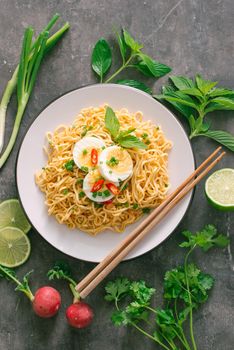 Dry instant noodles put egg with fresh herbs, garnish of cilantro and Asian basil, lemon, lime on dark stone background