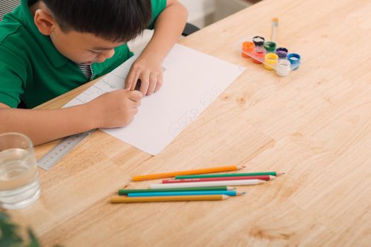 Cute confused smiling boy doing homework, coloring pages, writing and painting . Children paint. Kids draw. Preschooler with books in the library. Colorful pencils and paper on a desk. Creative boy.